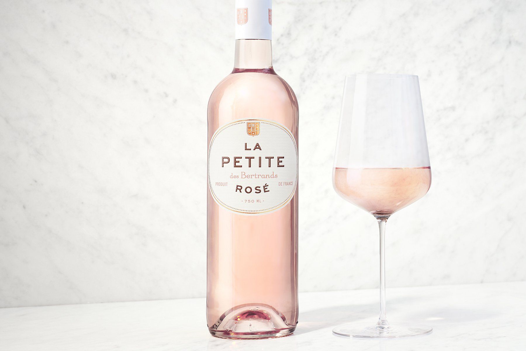 Bottle of La Petite Rosé wine next to a glass filled with the same pale pink wine. White background.