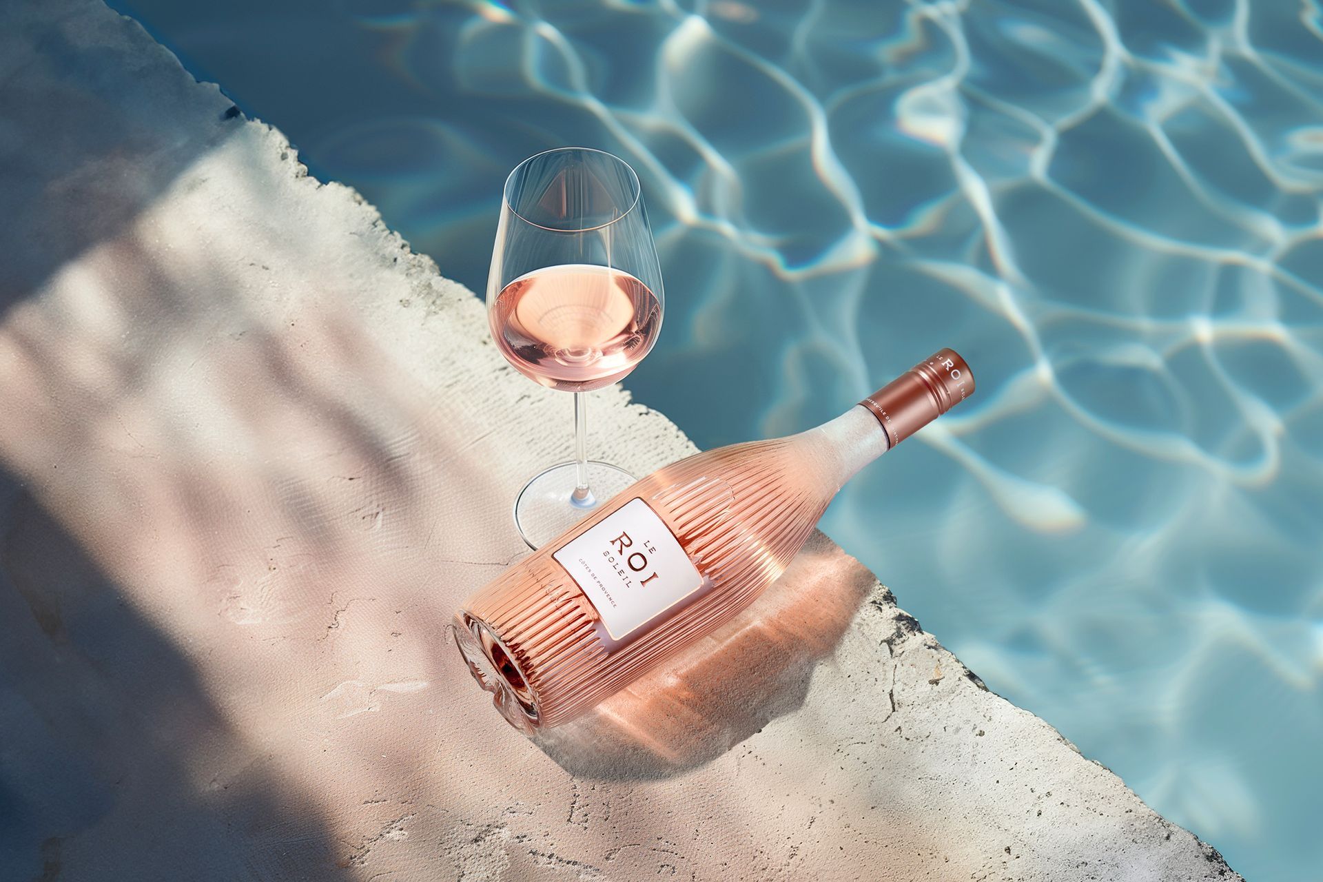 Rose wine bottle and glass on pool edge, with shimmering water.
