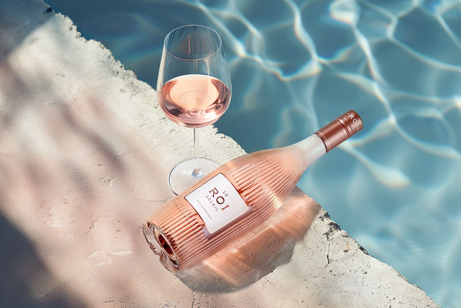 Rose wine bottle and glass poolside, reflecting in the water, sunlight.