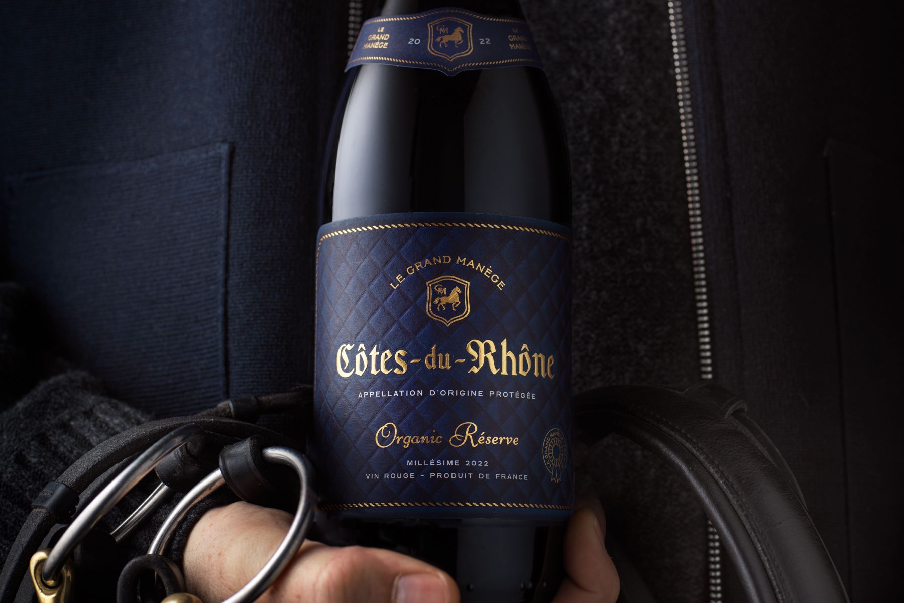 Person holding a dark blue Côtes-du-Rhône wine bottle with gold lettering; dark setting. Le Grand Manege