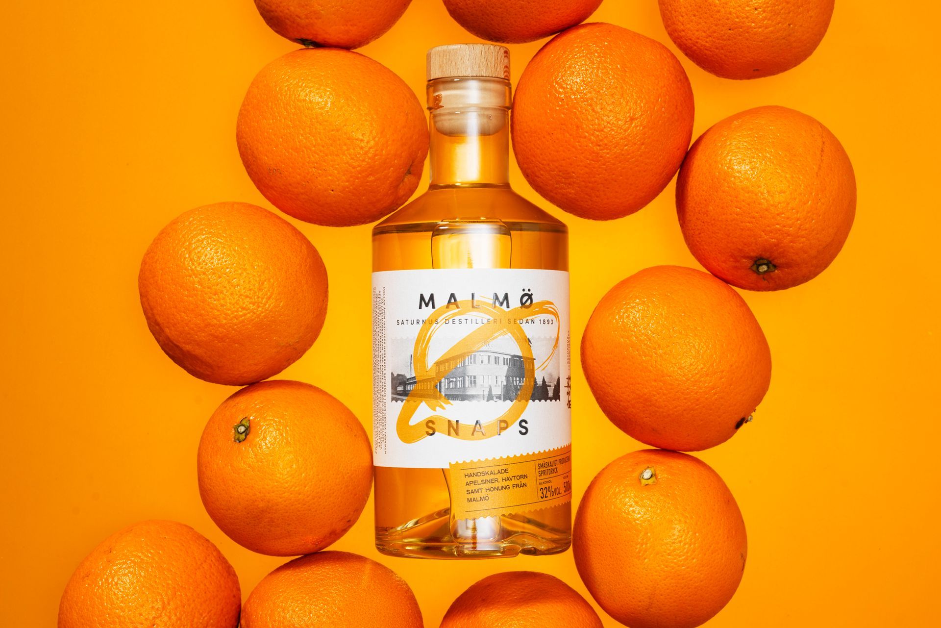 Bottle of orange-colored liquid, Malmö Snaps from Saturnus, surrounded by oranges on an orange background.