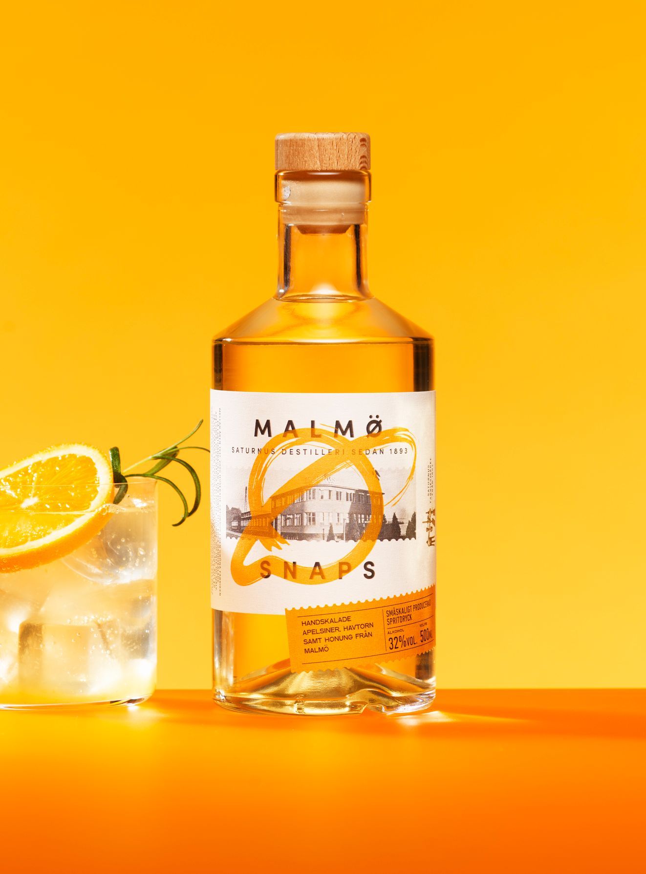 Bottle of Malmö Snaps with a cocktail, orange slice, and herbs against an orange background.
