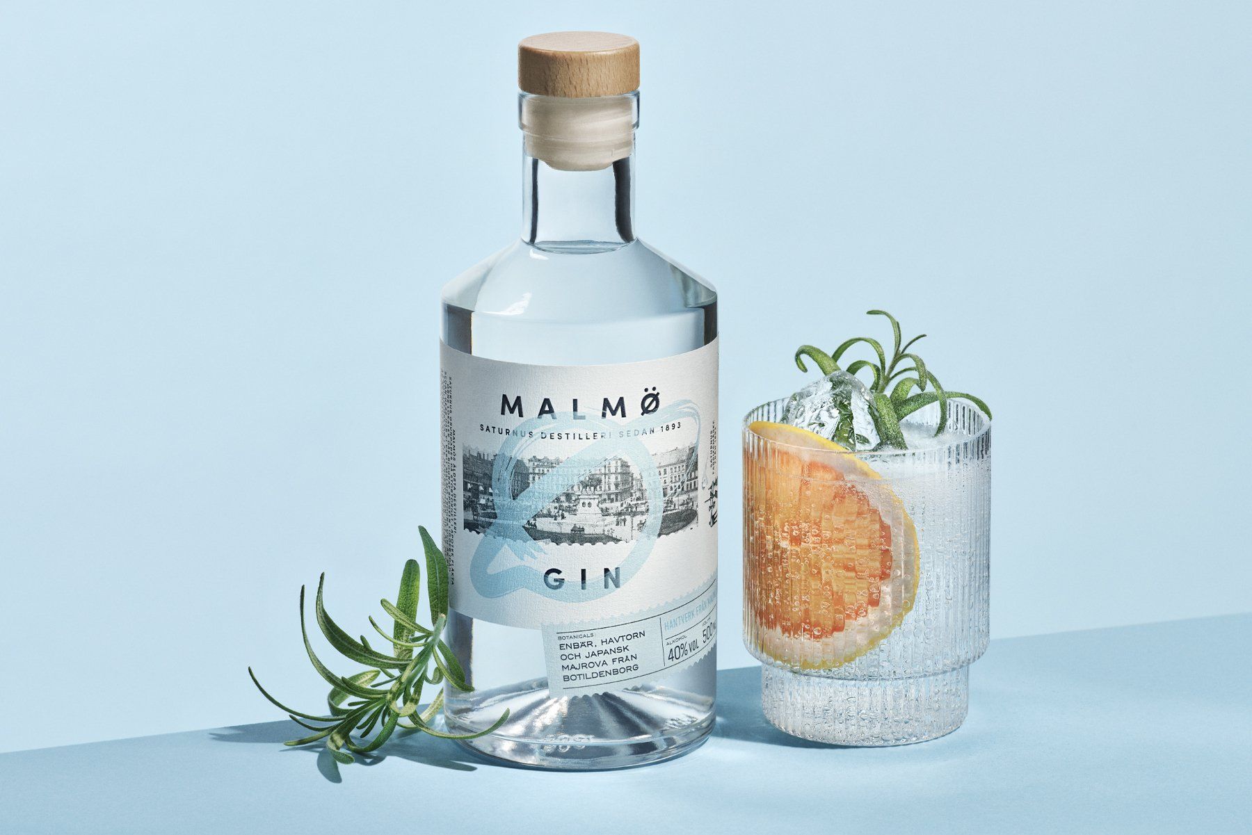 A bottle of Malmö Gin and a cocktail with grapefruit and rosemary on a blue surface.