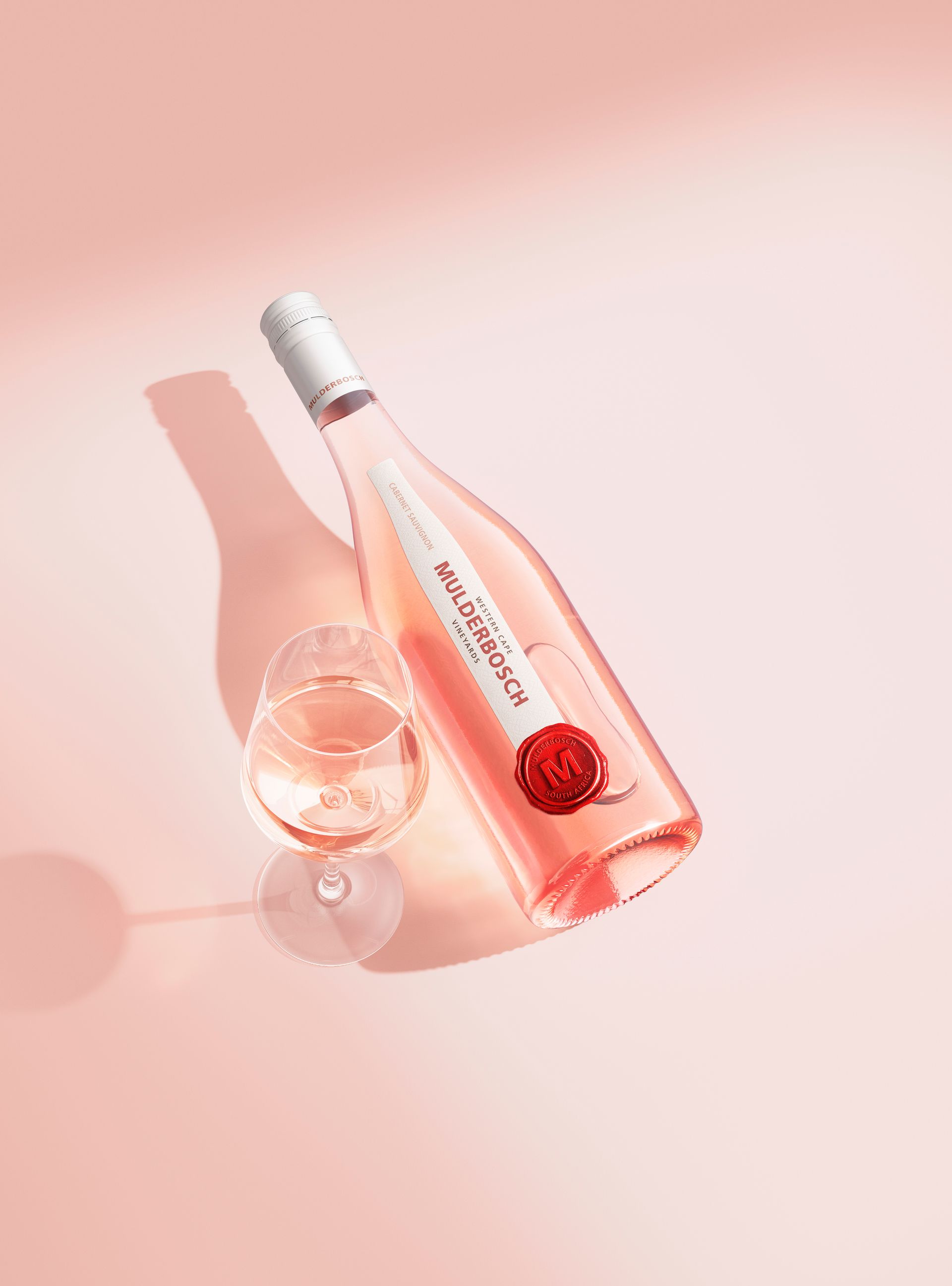 Bottle of rose wine with a filled glass on a pink surface; the bottle casts a shadow.