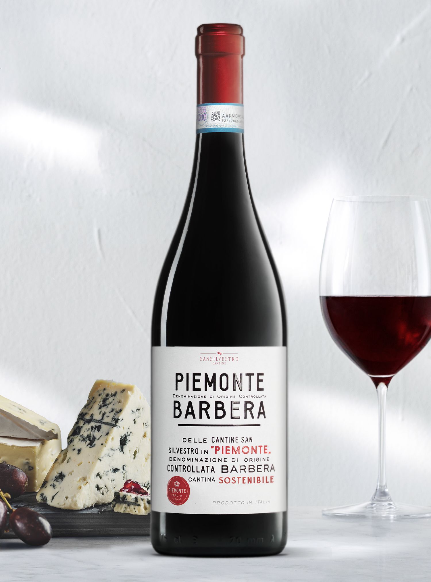 Bottle of Piedmont Barbera wine next to a glass of red wine and cheese on a white surface.
