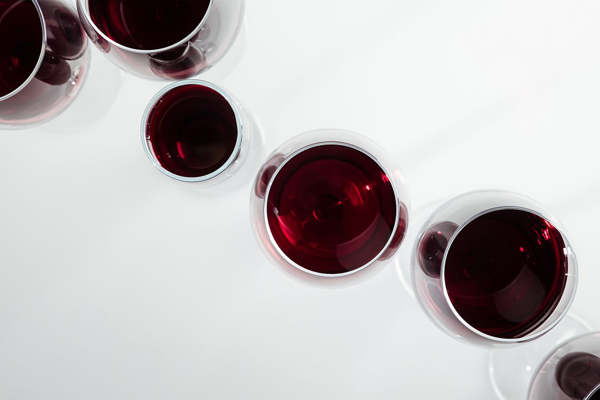 Six wine glasses filled with red wine, arranged on a white surface. Overhead view.
