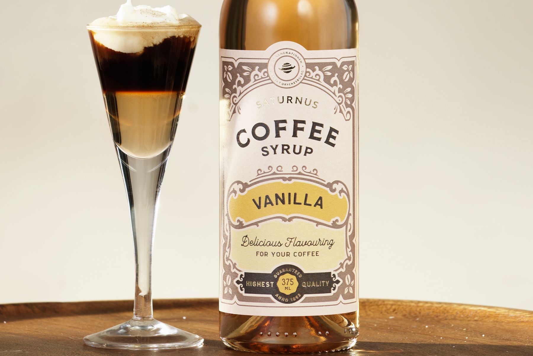 A layered coffee drink and vanilla coffee syrup bottle on a wooden surface from Saturnus.