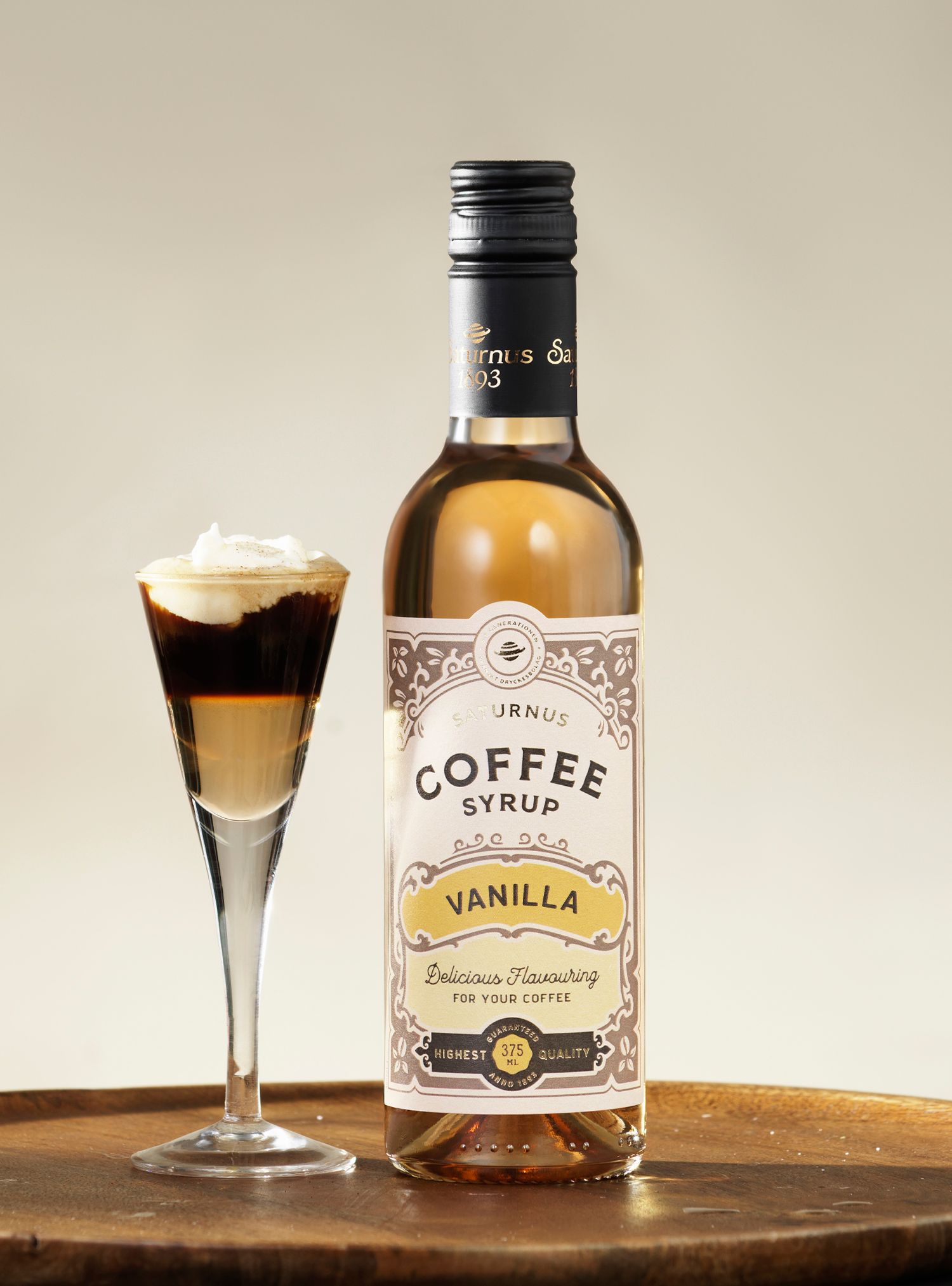 Bottle of vanilla coffee syrup next to a layered coffee drink in a slender glass with whipped cream.