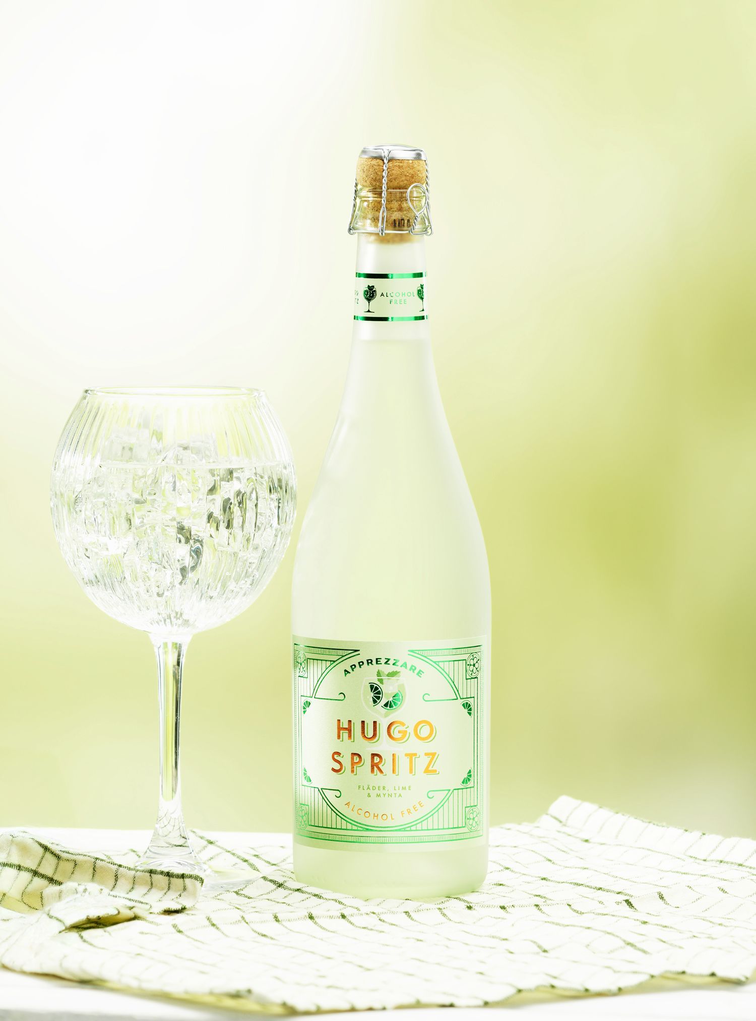 Bottle of Hugo Spritz, from Saturnus, and a filled wine glass on a white cloth, against a blurred green background.