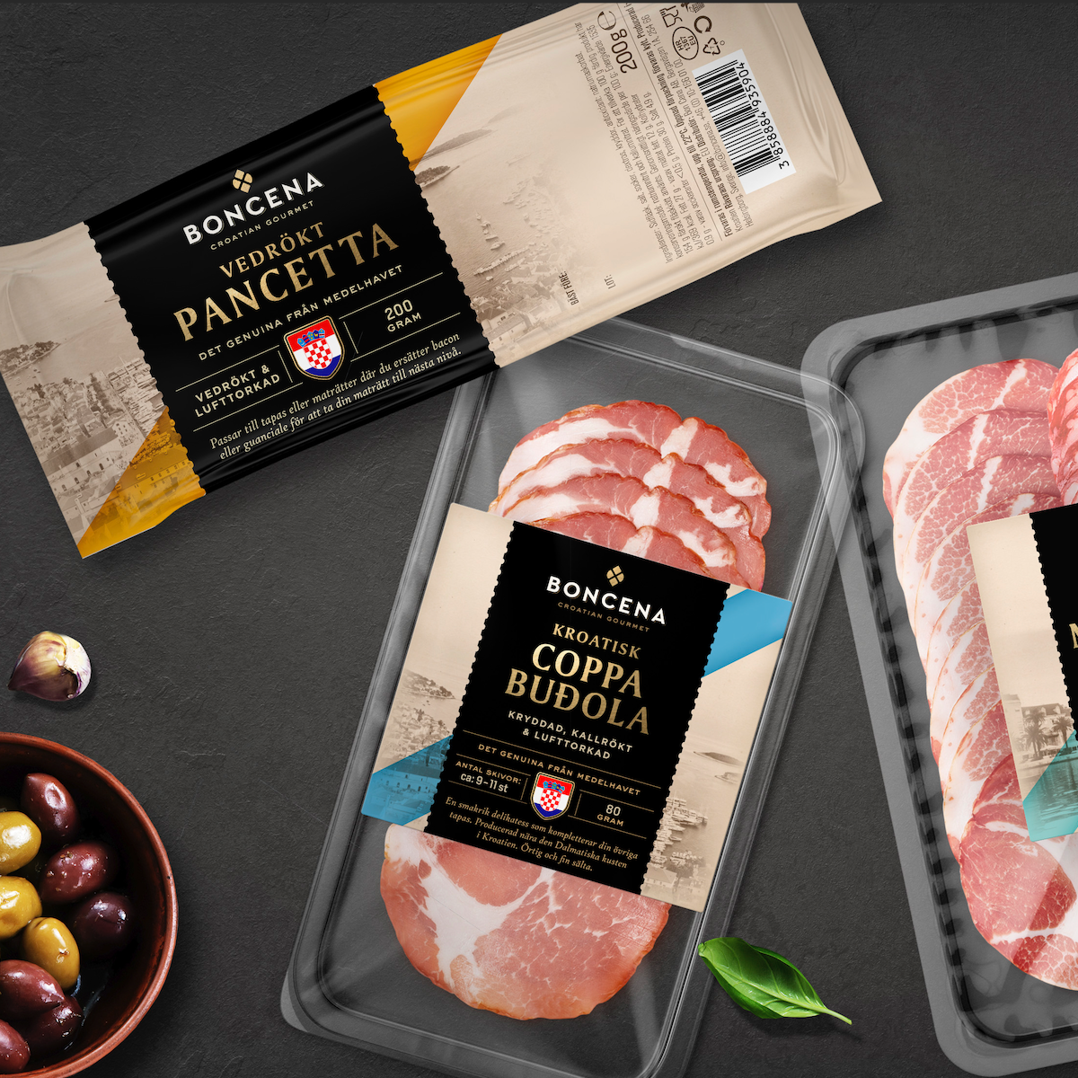 Packages of pancetta and coppa budola meat products arranged on a dark surface with olives.