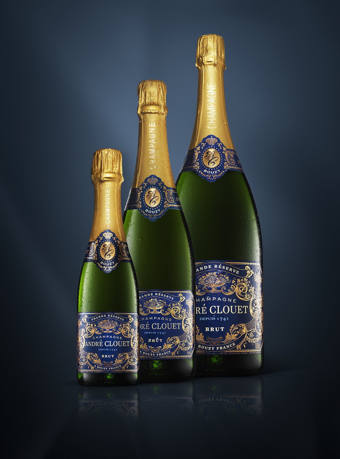 Three champagne bottles of varying sizes with gold foil tops and blue and gold labels on a dark background.