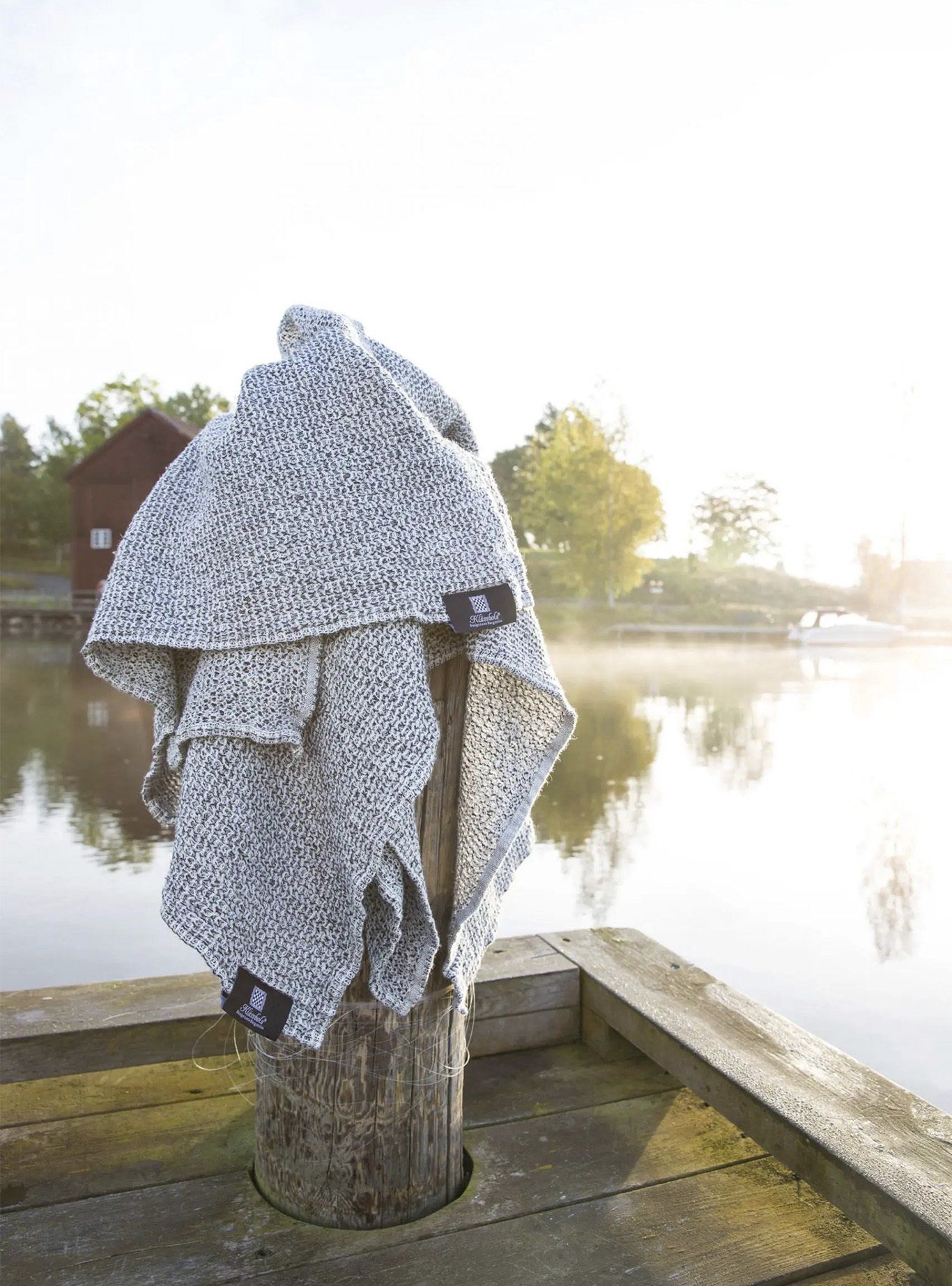 A textured gray blanket from Klässbols linneväveri draped over a wooden post by a calm lake in the morning sun.