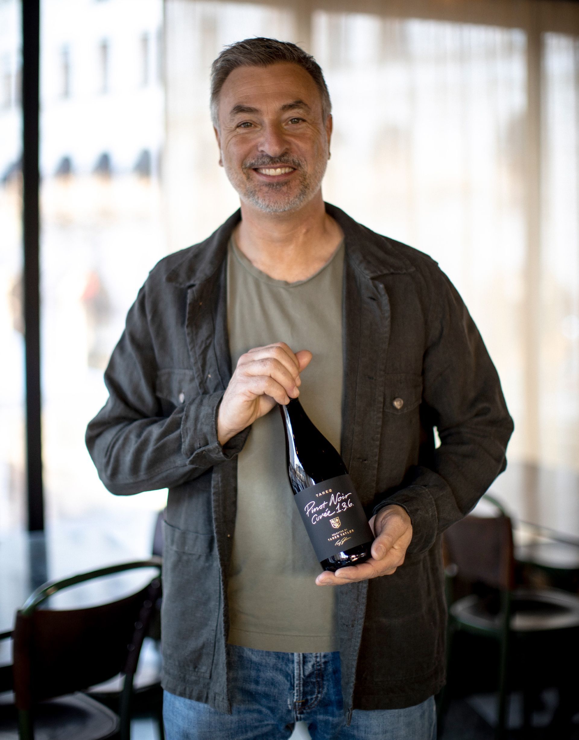 Tareq Taylor smiling, holding a bottle of wine. Indoors, wearing casual clothes.
