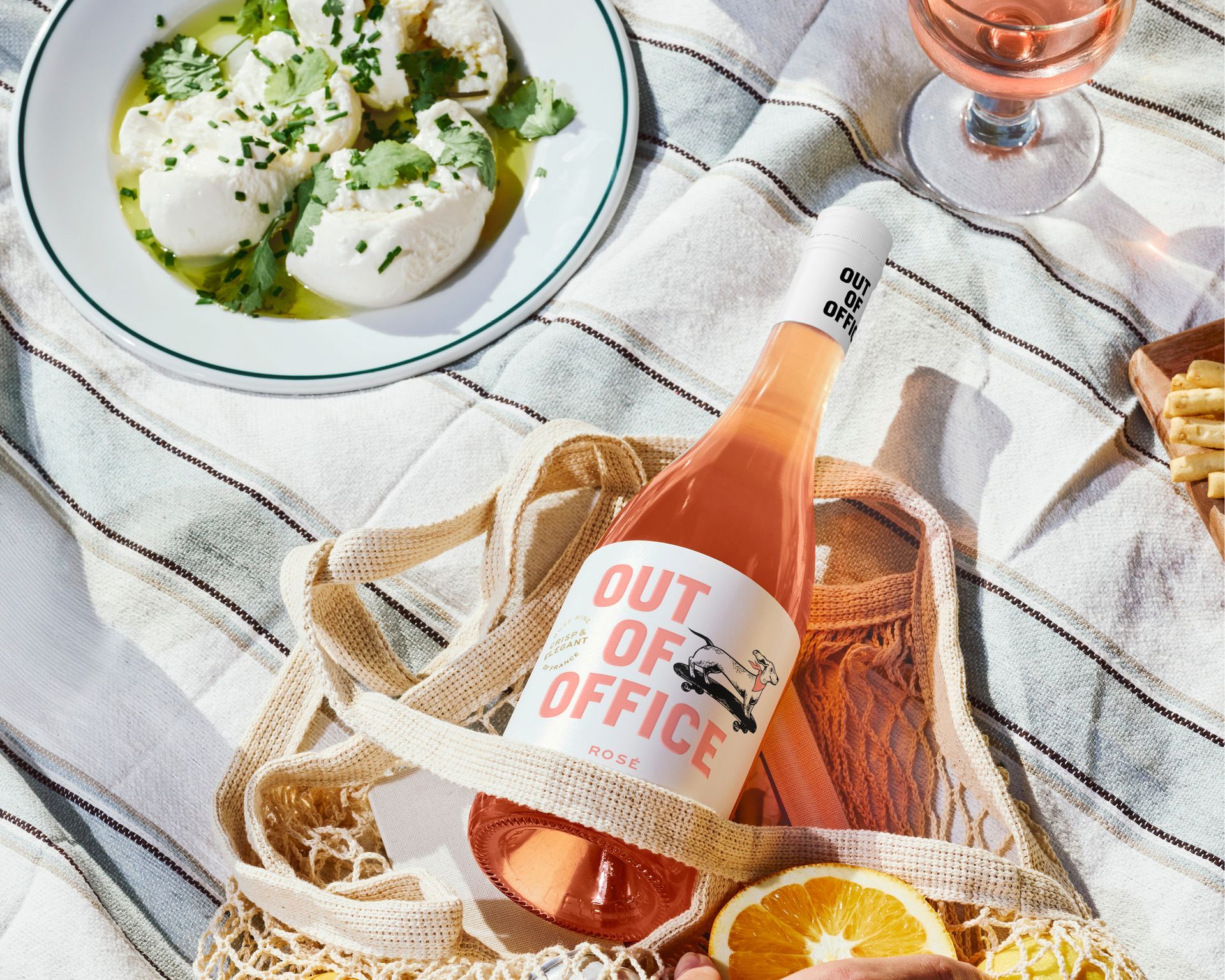 Rosé bottle labeled “OUT OF OFFICE” on a picnic blanket with plates of food and a wine glass
