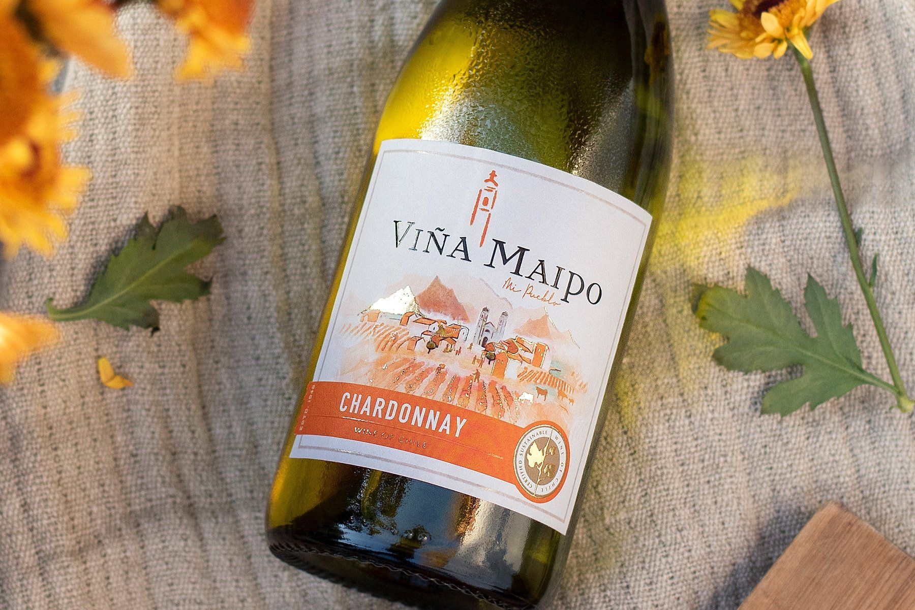 Bottle of Viña Maipo Chardonnay wine from VCT on a light fabric, with yellow flowers and green leaves.