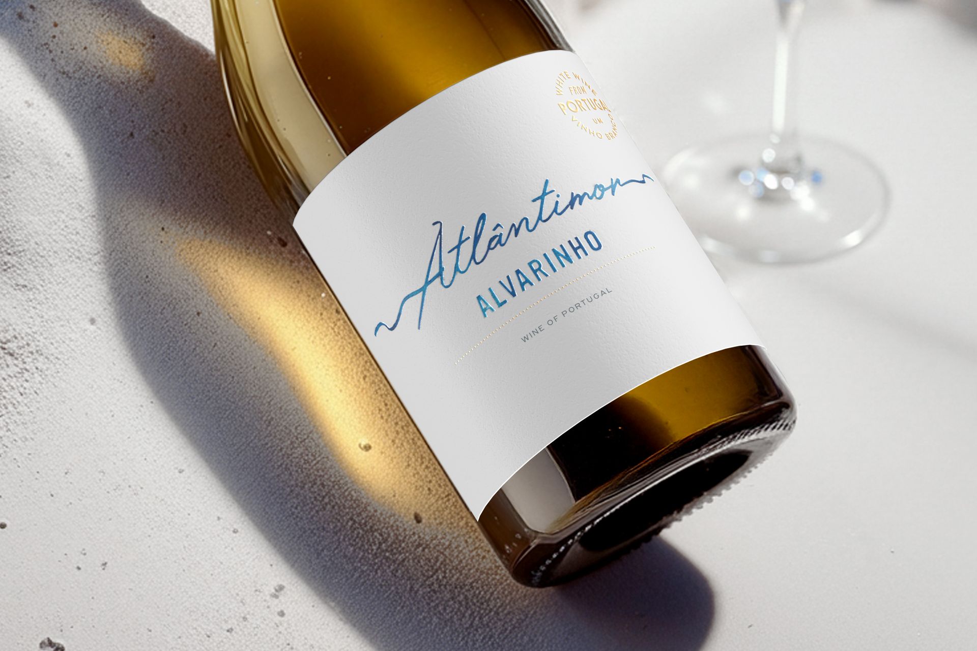 Bottle of Atlântico Alvarinho white wine, blue and gold label, angled on a white surface with a shadow.