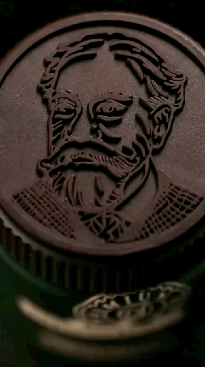 Dark brown cap with an embossed portrait of a man with a mustache and goatee, atop a green bottle.