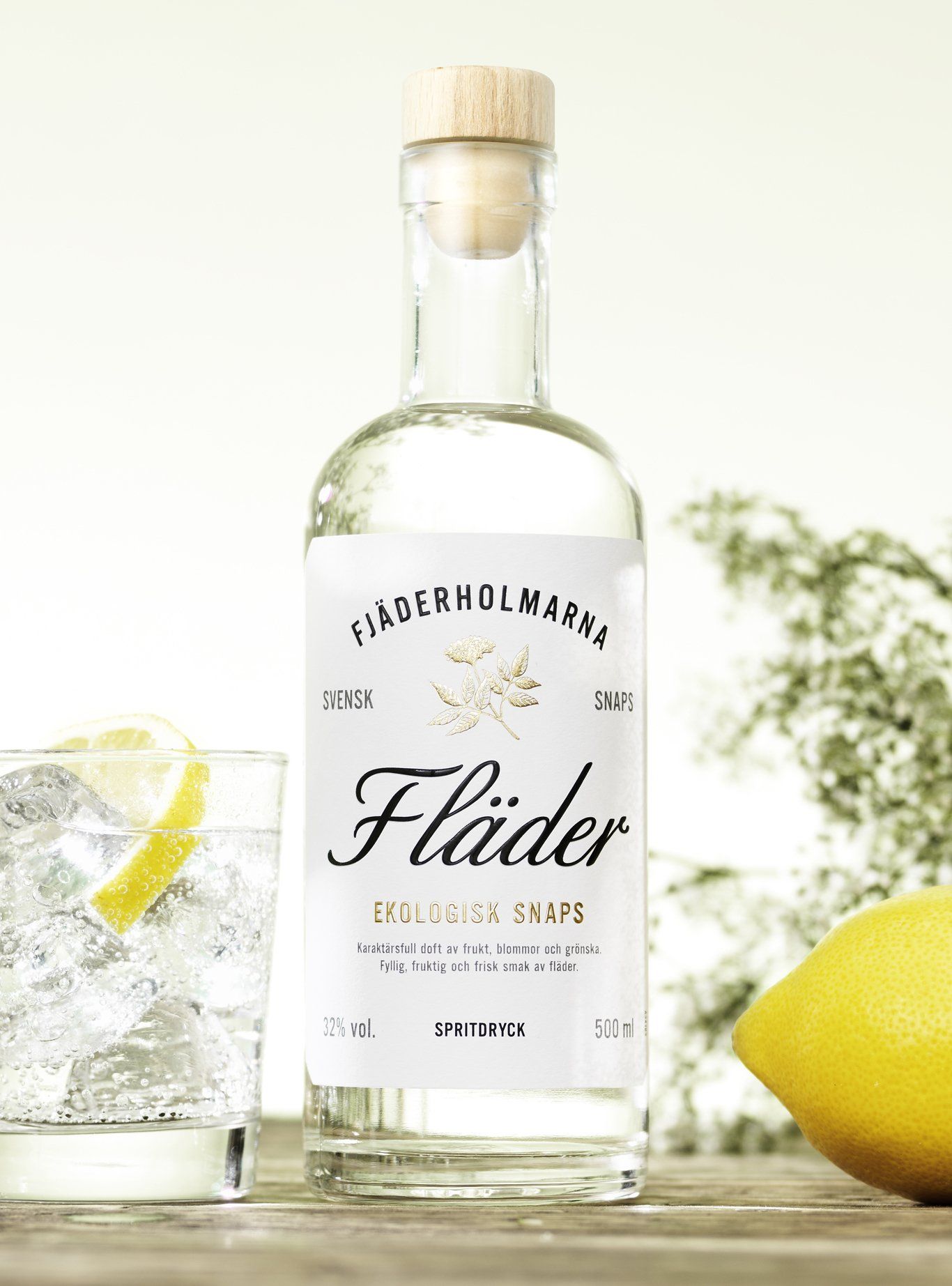 Bottle of Fläder liqueur next to a glass with a lemon slice and a lemon on a wooden surface.