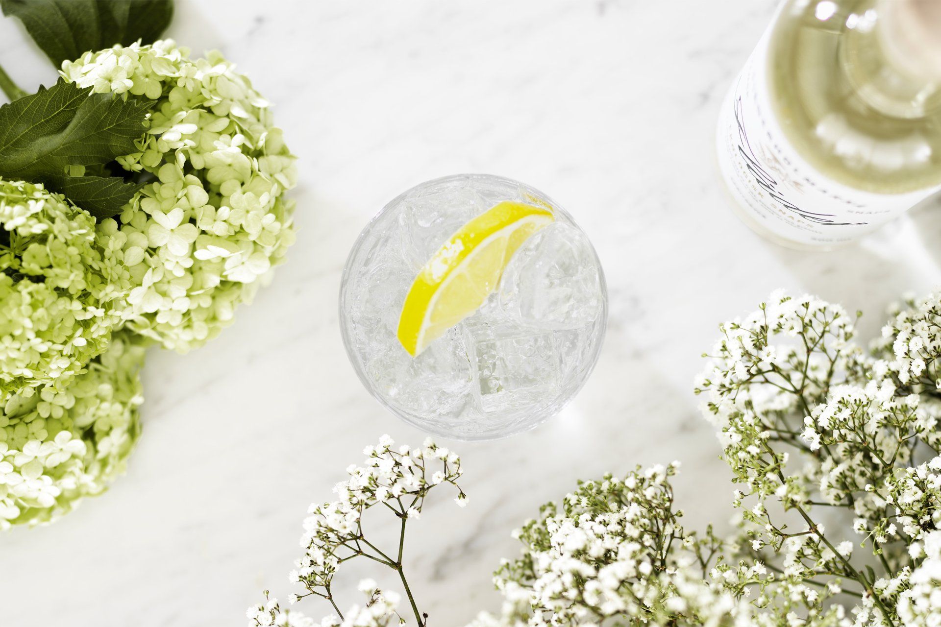 Gin and tonic in a glass with a lime wedge, surrounded by flowers and a gin bottle, on a white surface.
