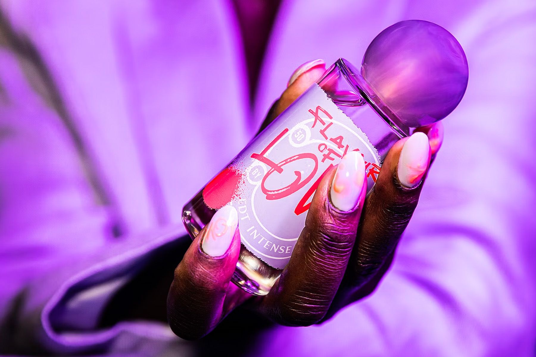 Hand holding a perfume bottle, lit with purple light. Bottle label reads 