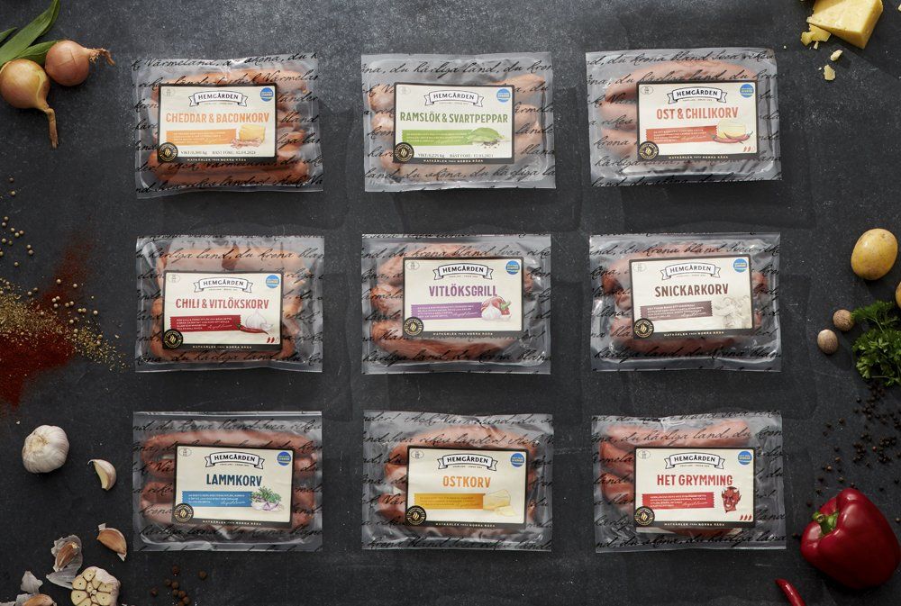 Nine sausage packages, various flavors, arranged on a dark surface with ingredients like onions and spices from Hemgården.