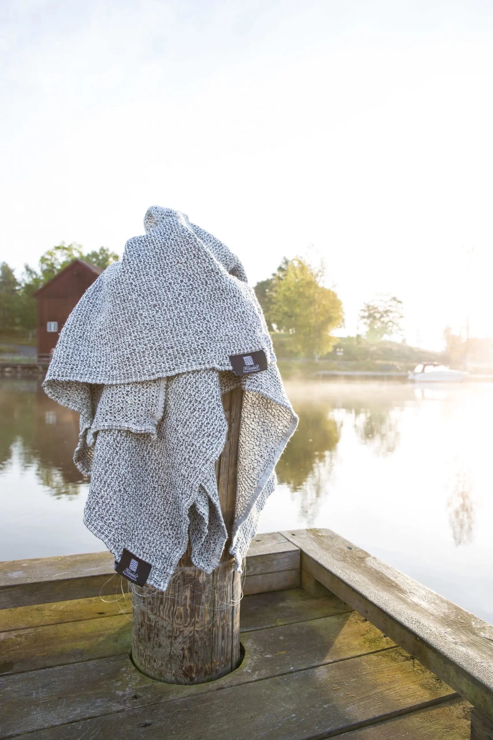 A speckled blanket draped over a wooden post on a pier, with a lake and sunlit sky in the background.