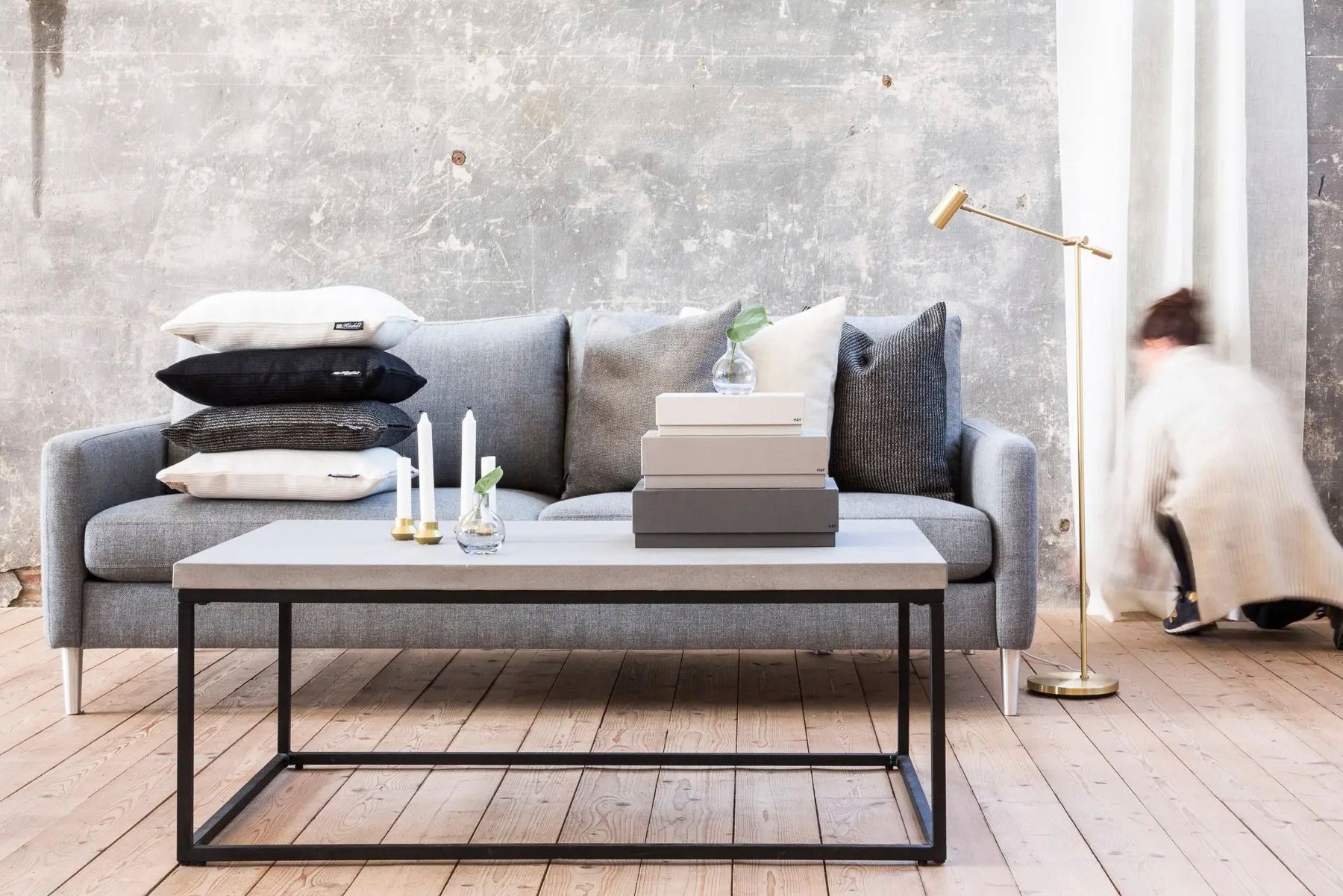 Gray couch with pillows, concrete coffee table, person adjusting curtain.