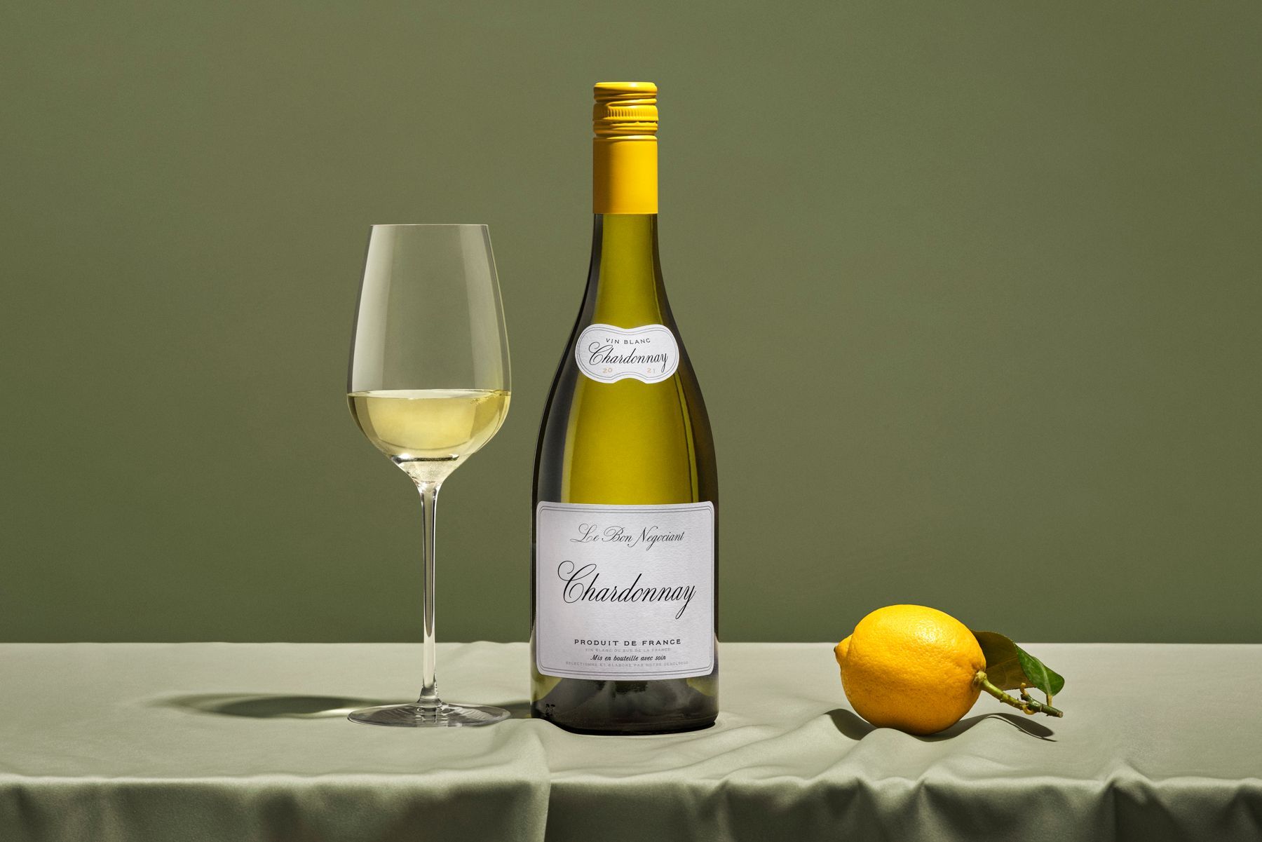 A bottle of Chardonnay with a full wine glass and a lemon on a green-draped table.