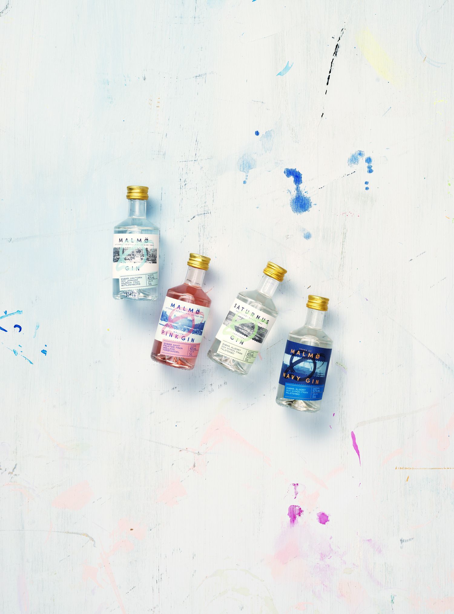 Four small, colorful bottles with gold lids on a splattered white surface.