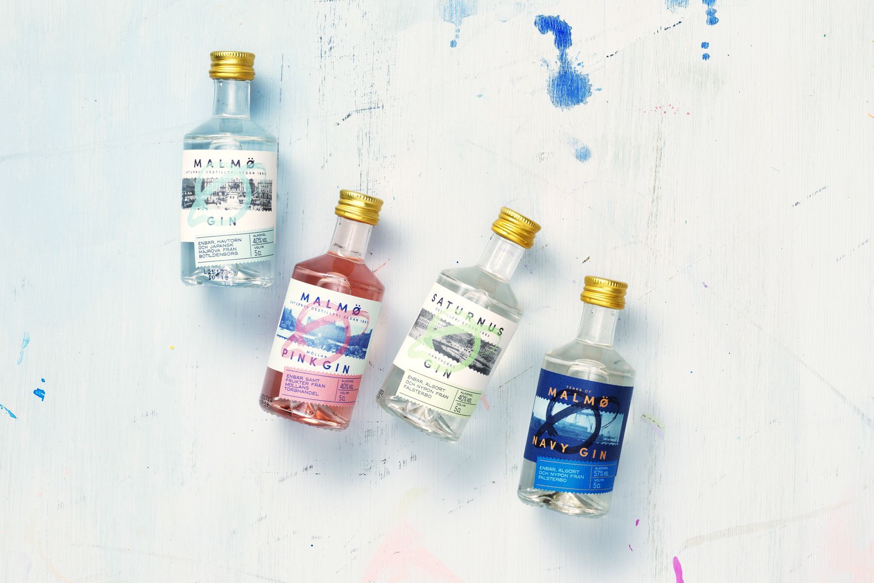 Four miniature liquor bottles, Saturnus Malmö Gin, with colorful labels on a splattered white surface.