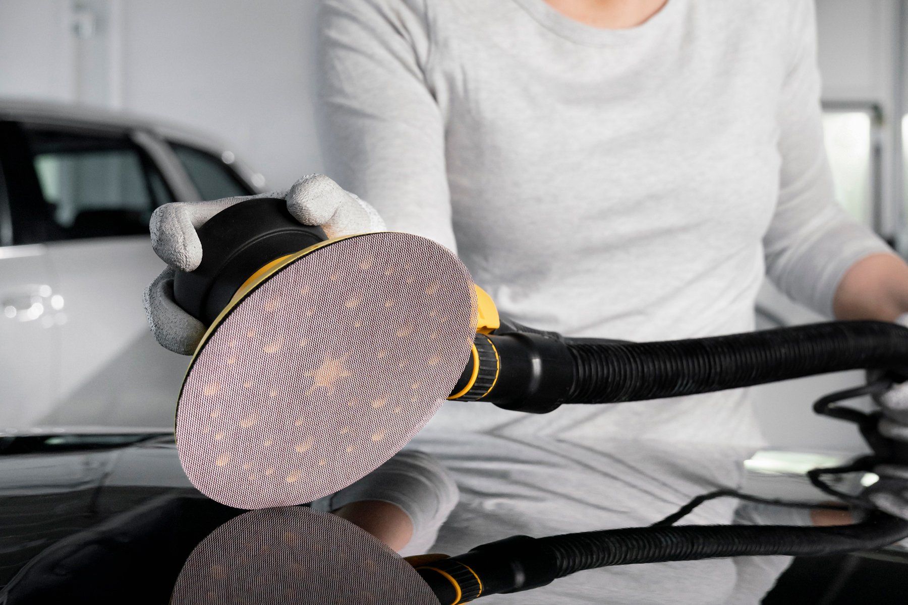 Person using a sander on a car's glossy black surface, wearing gloves, in a garage.