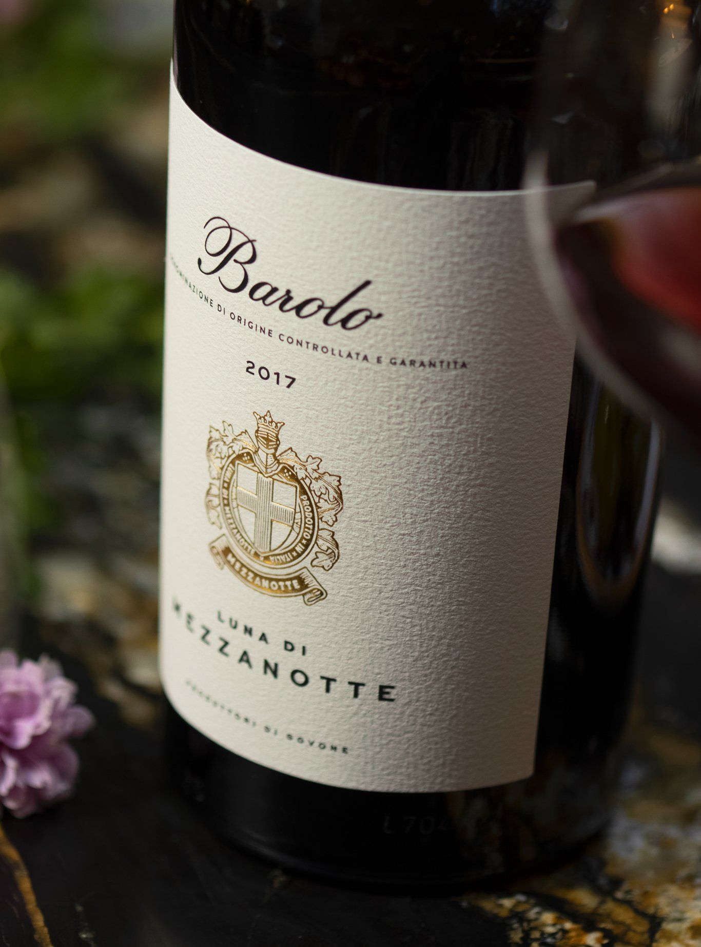 Bottle with a Barolo label, partially obscured by a wine glass. Label has gold and white details, with year 2017 visible.