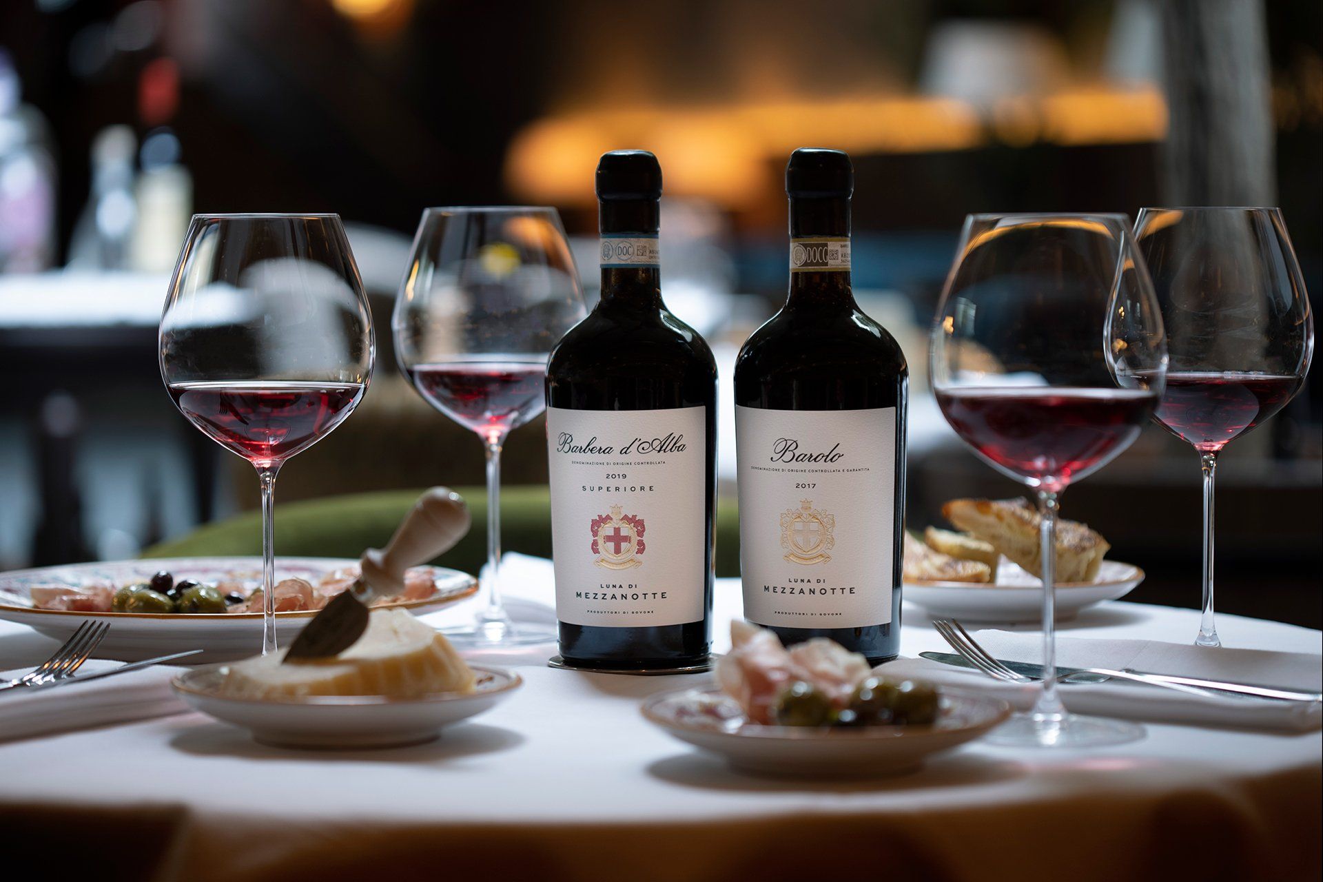 Table setting with two wine bottles, glasses filled with red wine, cheese, and olives.