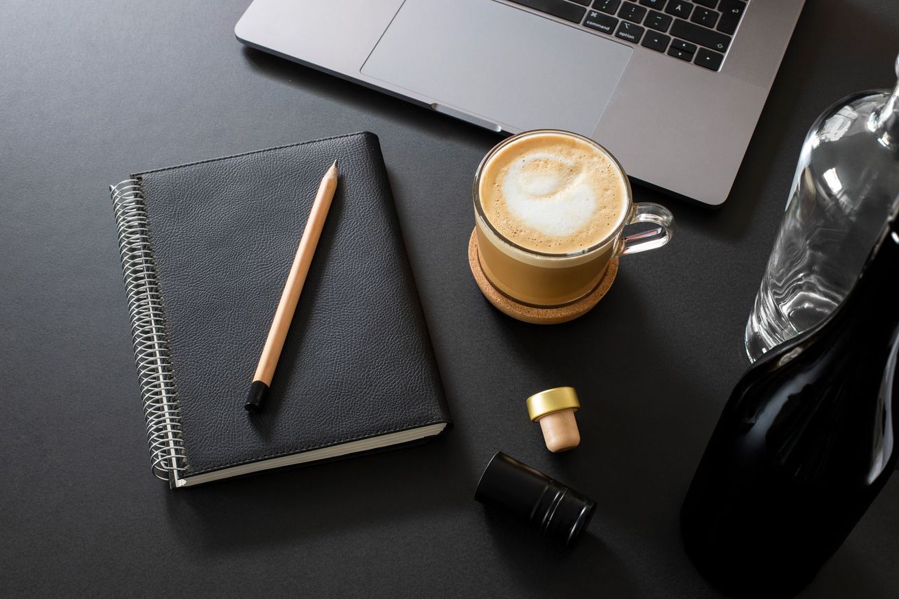 Notebook, pencil, laptop, coffee, and wine bottle on a dark surface.
