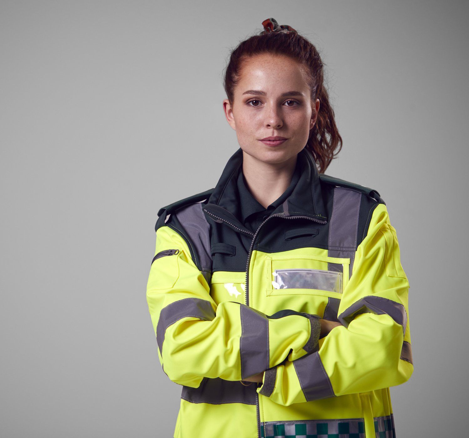 Paramedic in a yellow jacket with arms crossed, standing in front of a gray backdrop.