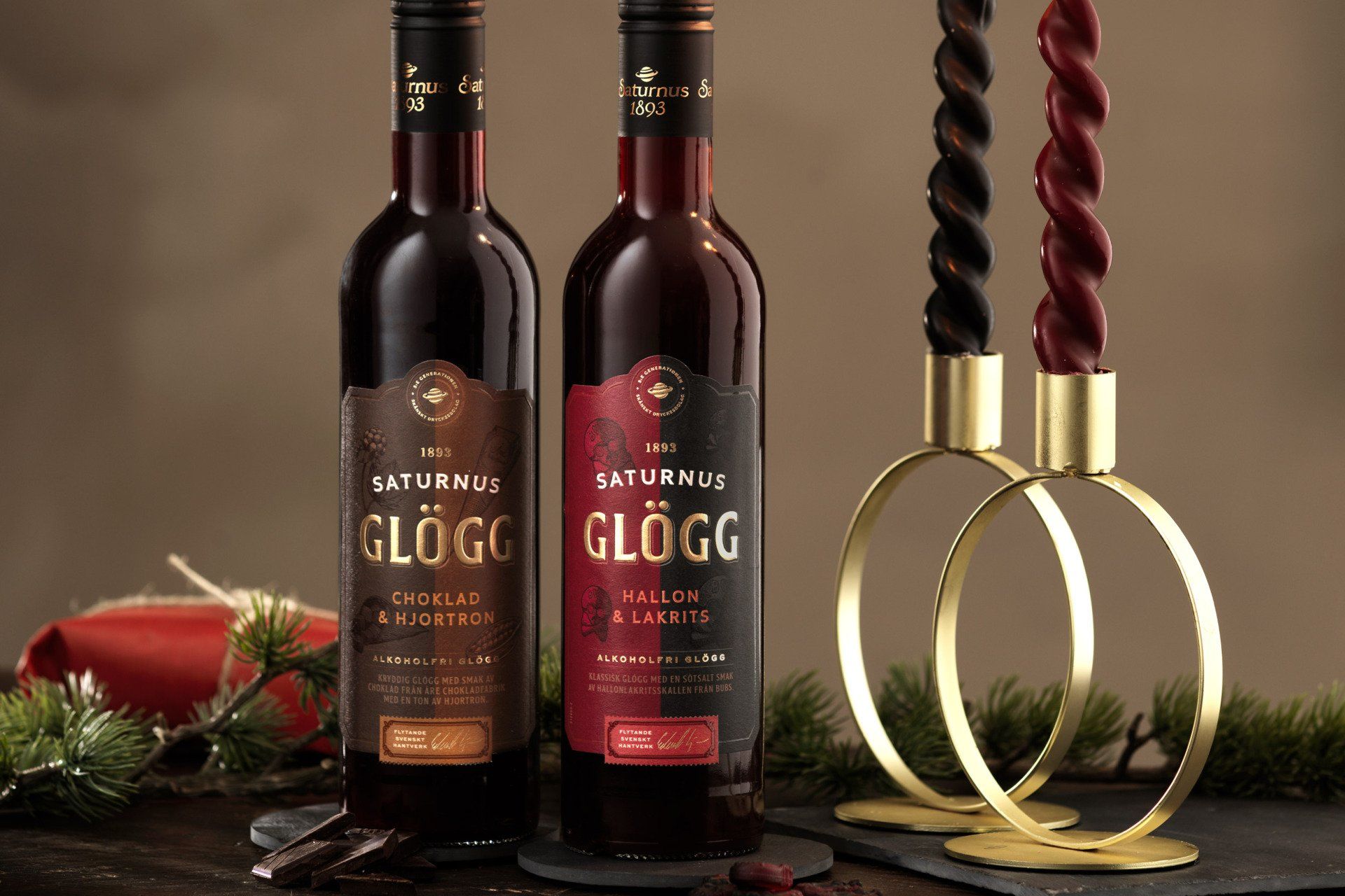 Two bottles of Glögg, one chocolate, one spiced, next to festive candles and greenery.