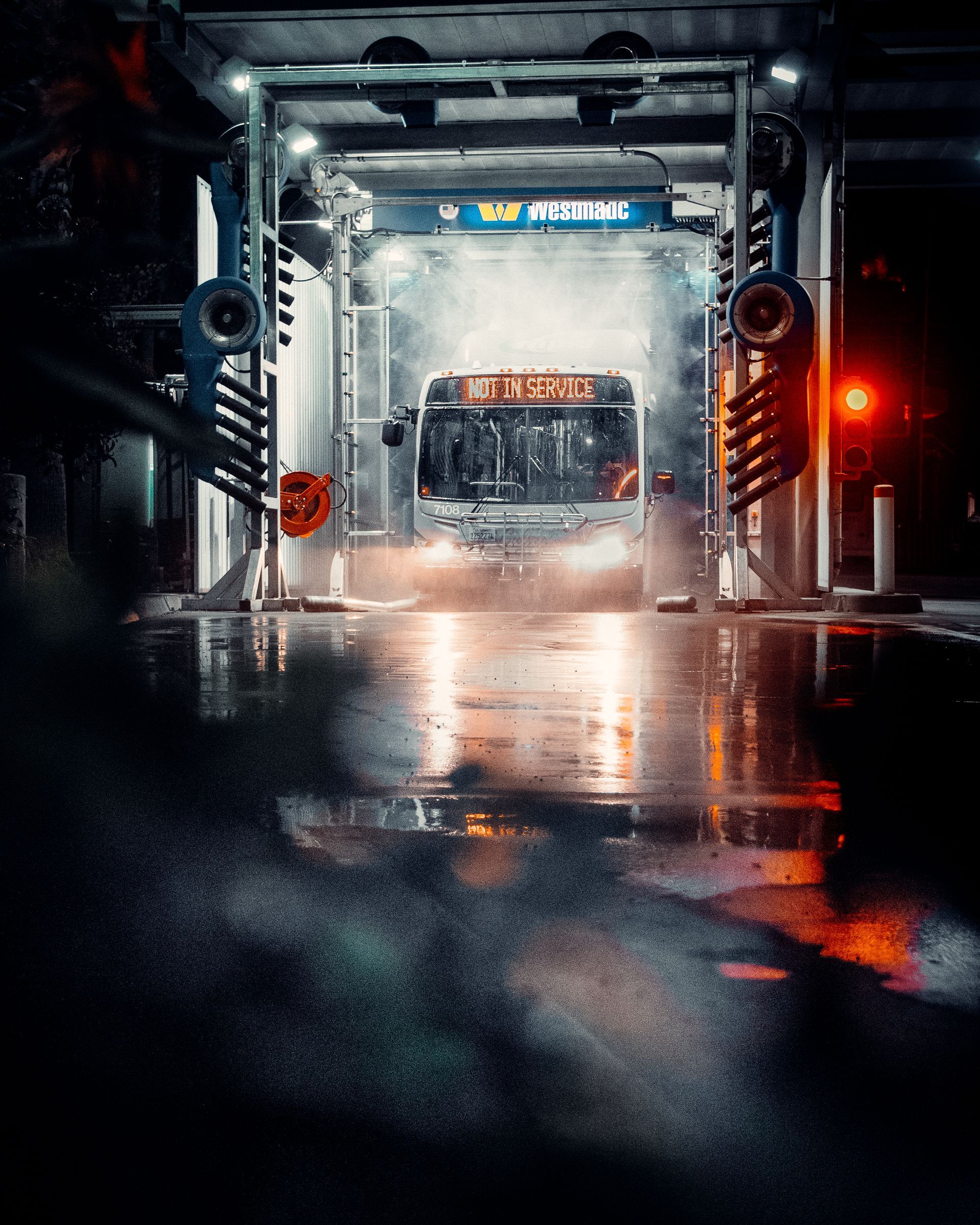 Bus driving through a car wash, illuminated by bright lights, creating mist and reflections on wet ground.