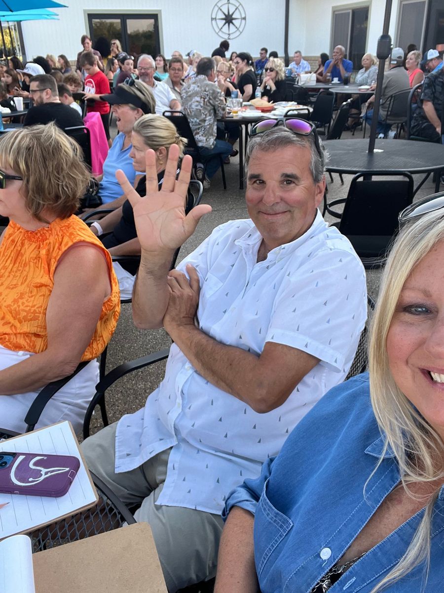 A group of people are sitting at tables outside and one man is waving