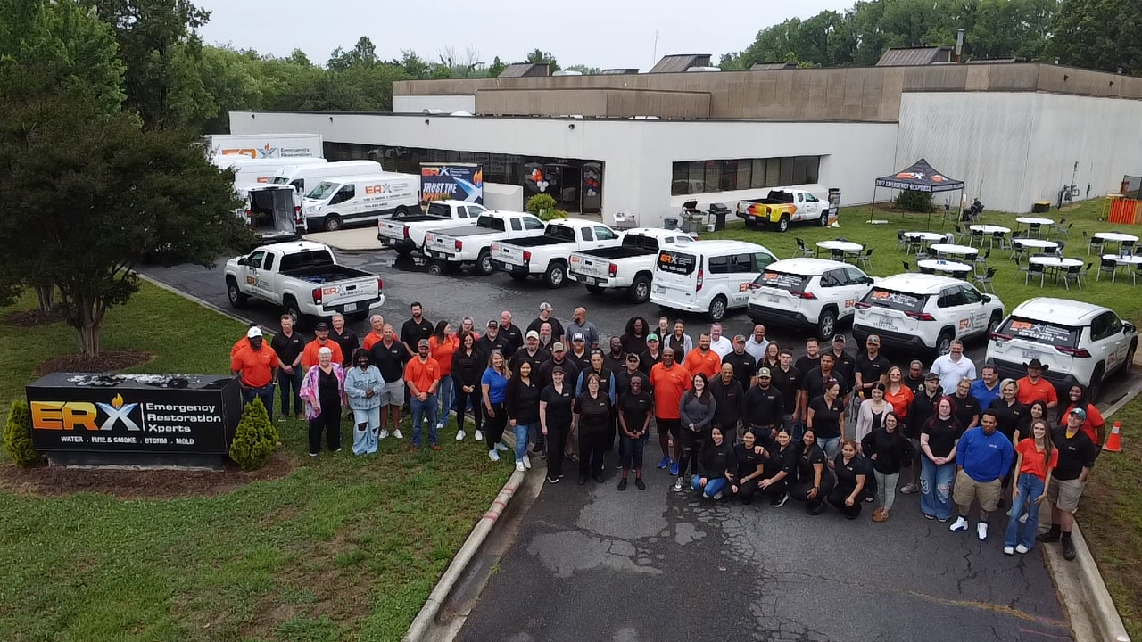 Large group of volunteers posing outside trucks and vans in a parking lot beside a beige building.