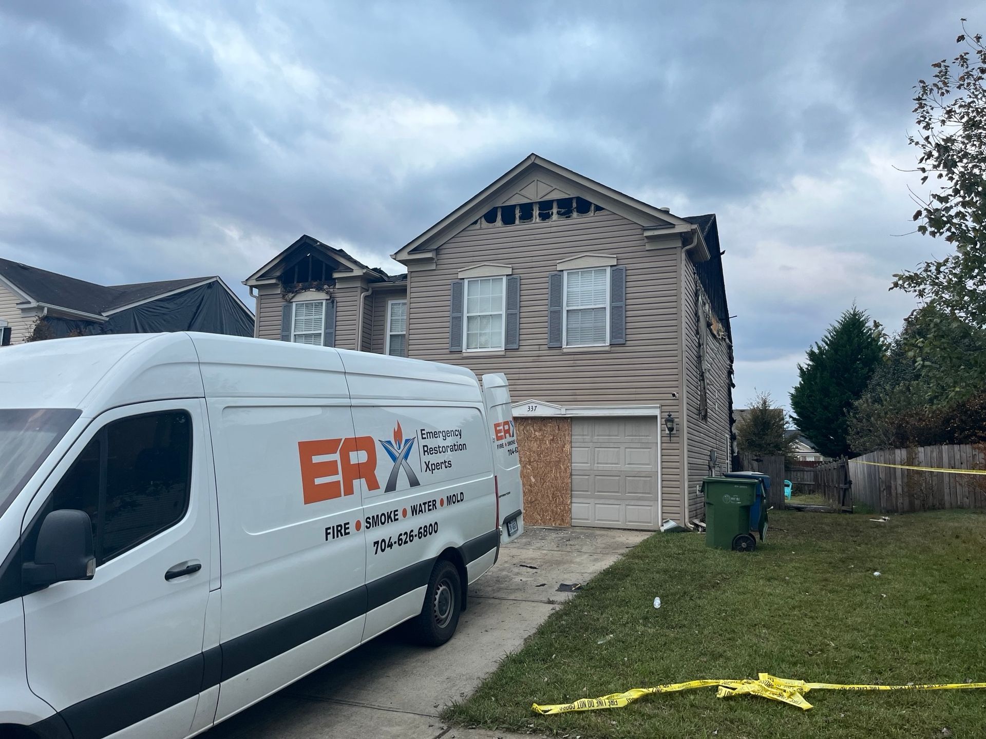 White service van parked by a two-story beige house with boarded garage and yellow caution tape in the yard