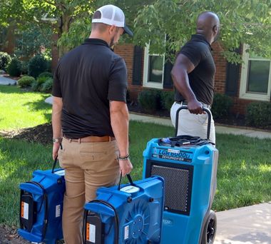 Two men carry blue portable equipment on a sidewalk in a sunny residential area