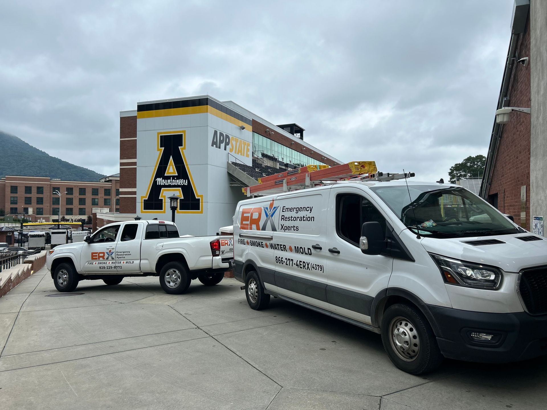 Two white work trucks parked on a street beside a building with an Appalachian State logo.
