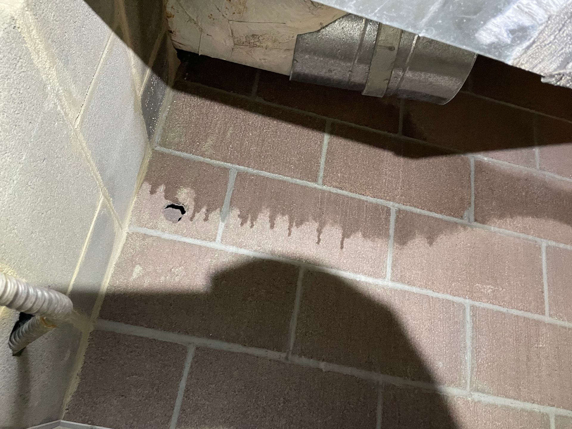Close-up of a brick floor beside a wall, with a metal duct and a shadow across the scene.