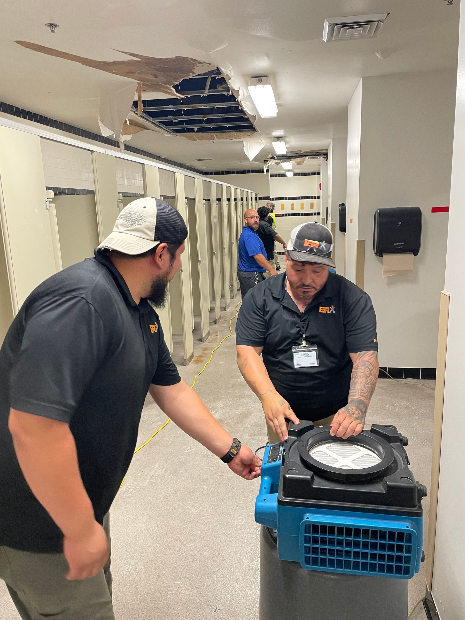 Two workers move equipment through a hallway with a damaged ceiling panel.