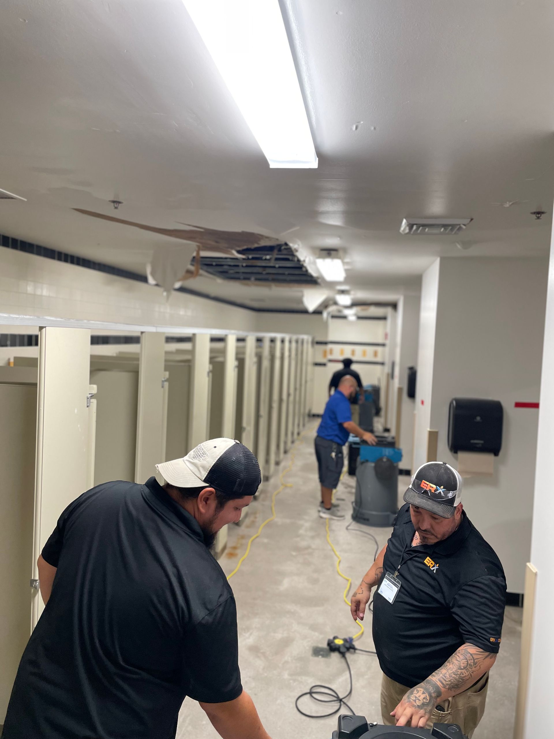 Workers cleaning a tiled hallway with buckets, cords, and supplies under bright fluorescent lights