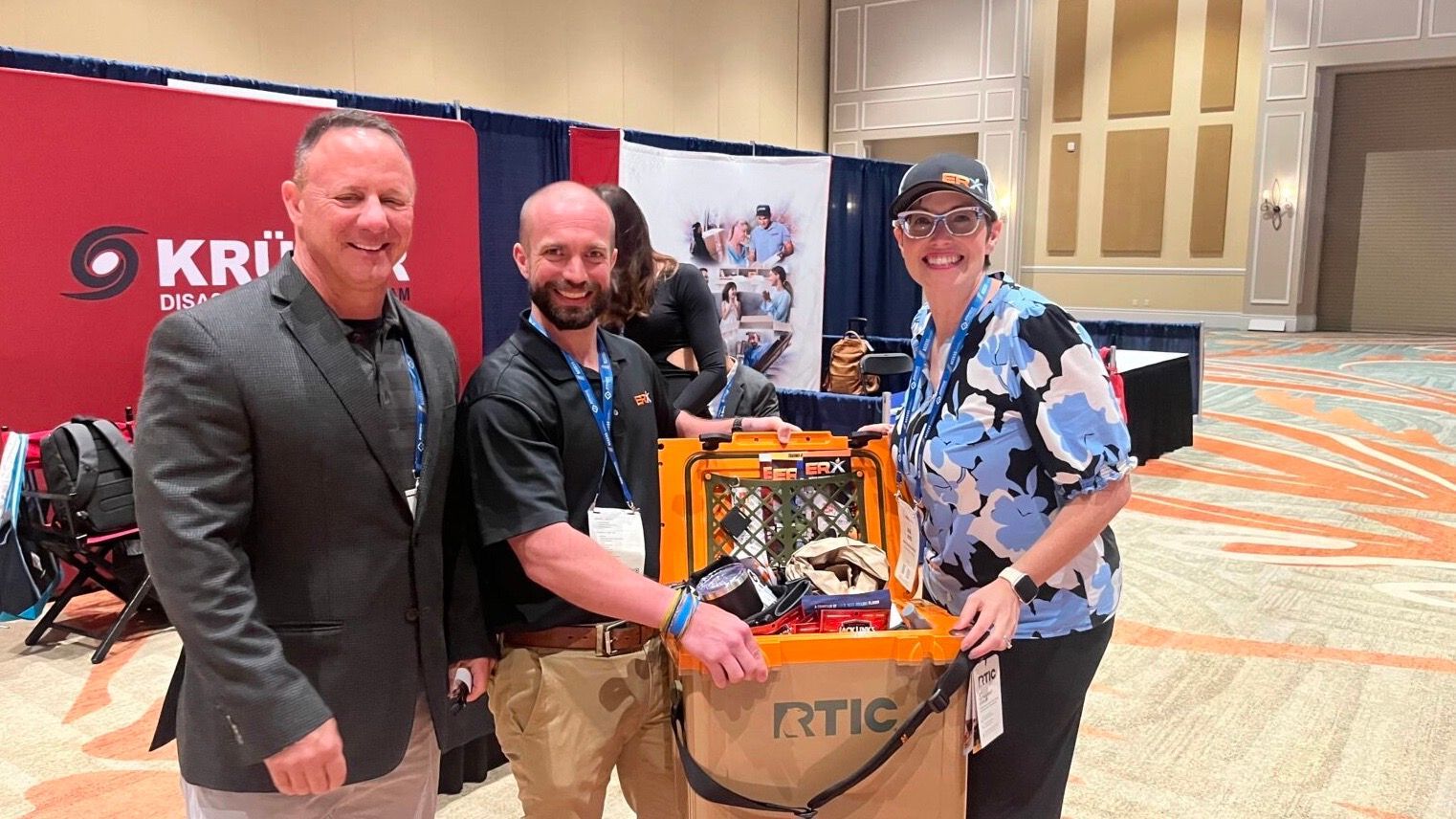Three men posing with an orange robotics platform at a conference booth, smiling in a convention hall