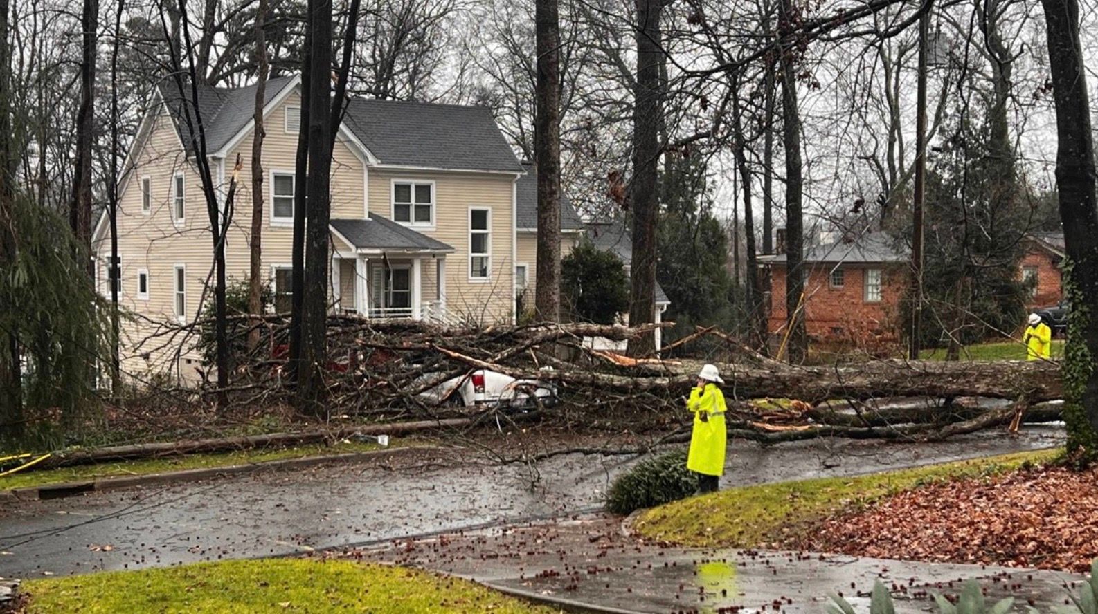 Storm damage cleanup on a rainy residential street, with fallen trees, debris, and workers in neon jackets