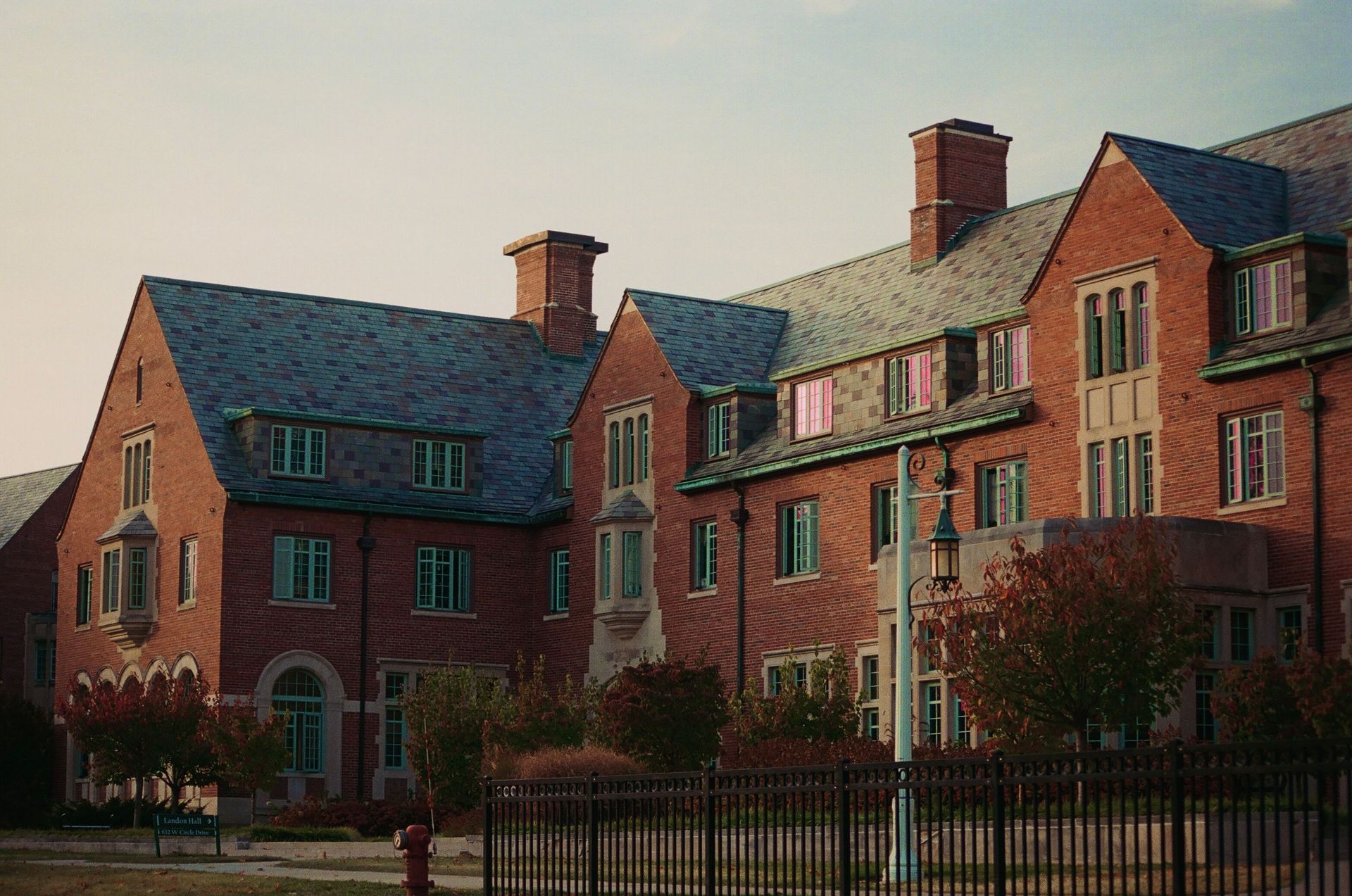 Red brick campus building with gabled roofs, chimneys, and trees behind a black fence.