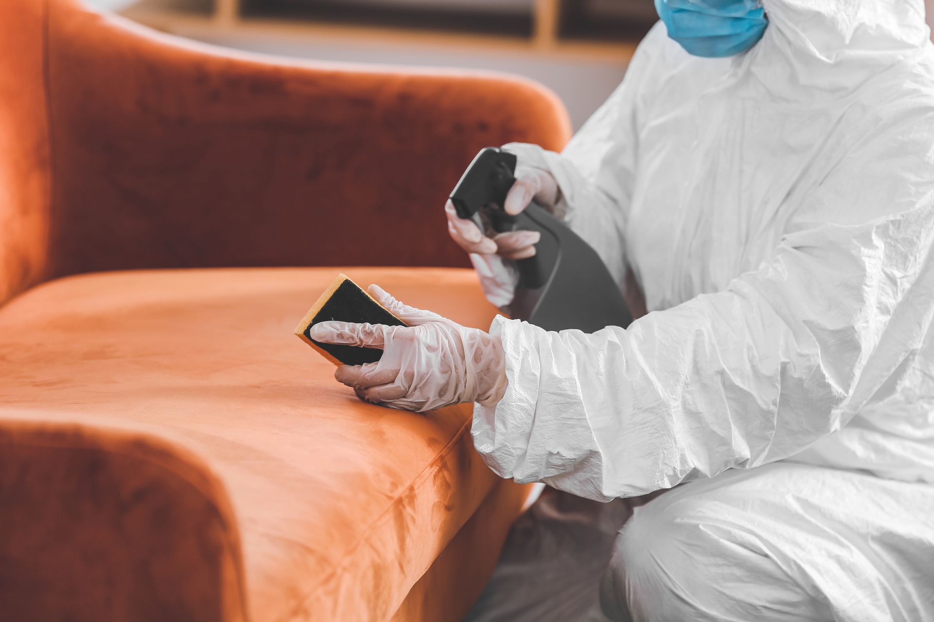 Worker in protective suit sanitizing an orange couch with disinfectant spray