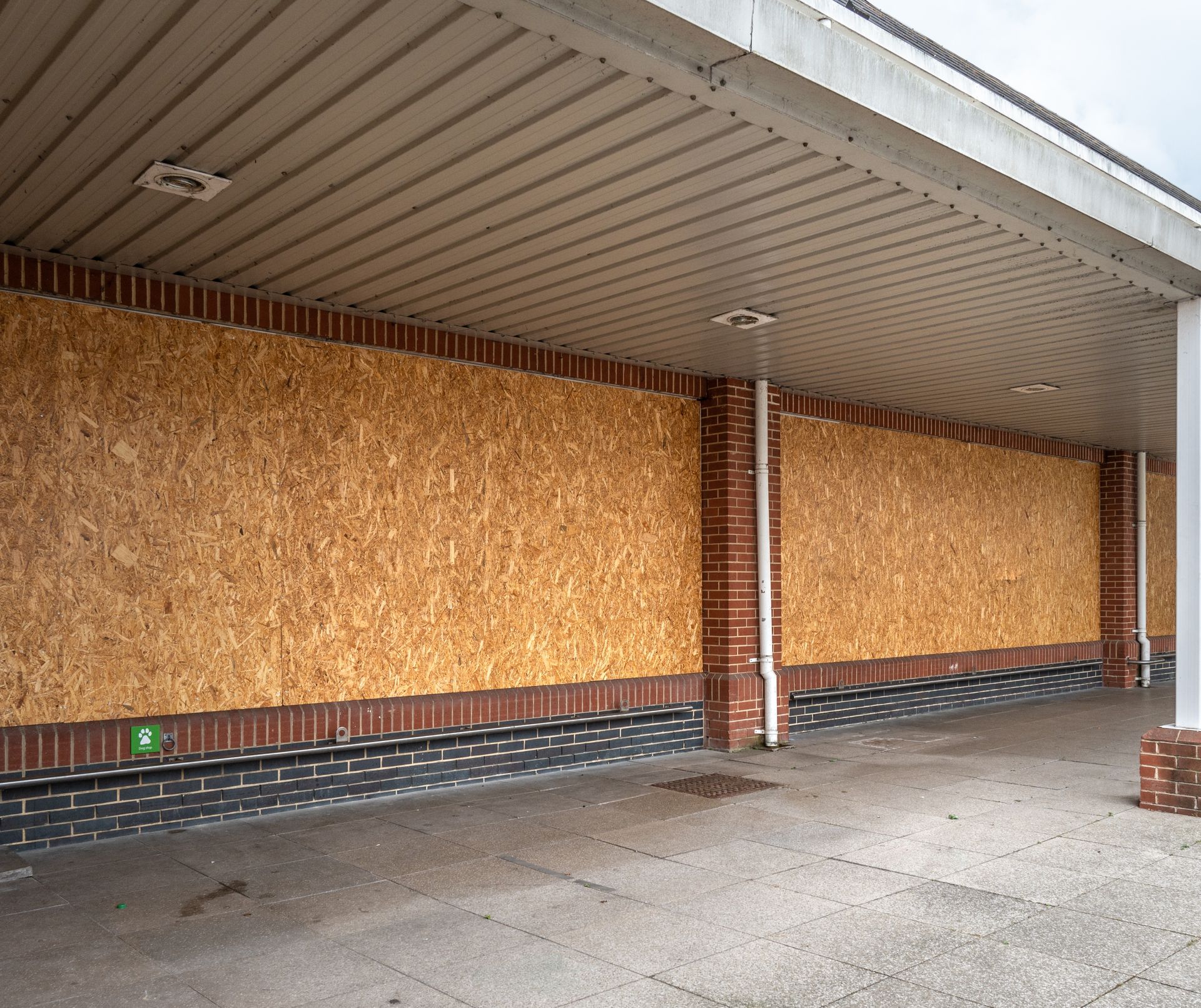 Boarded-up storefront with plywood windows, brick base, and a covered concrete walkway.